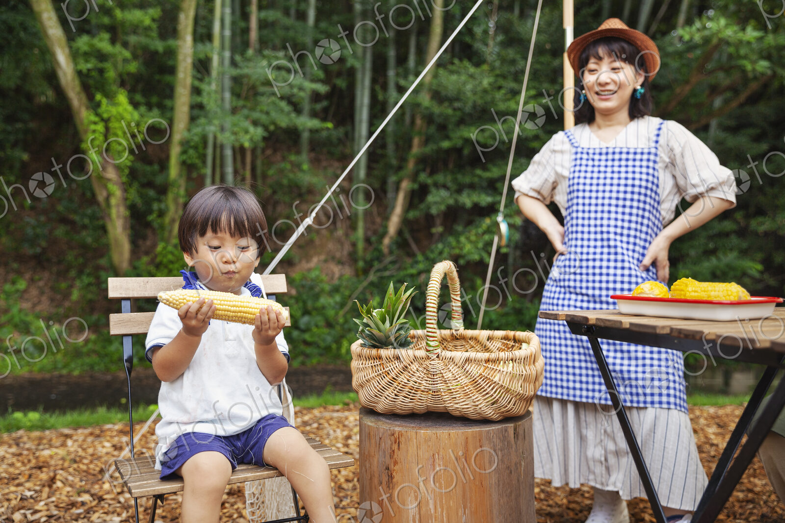 Japanese woman standing outdoors, wearing hat and apron and young boy sitting on…