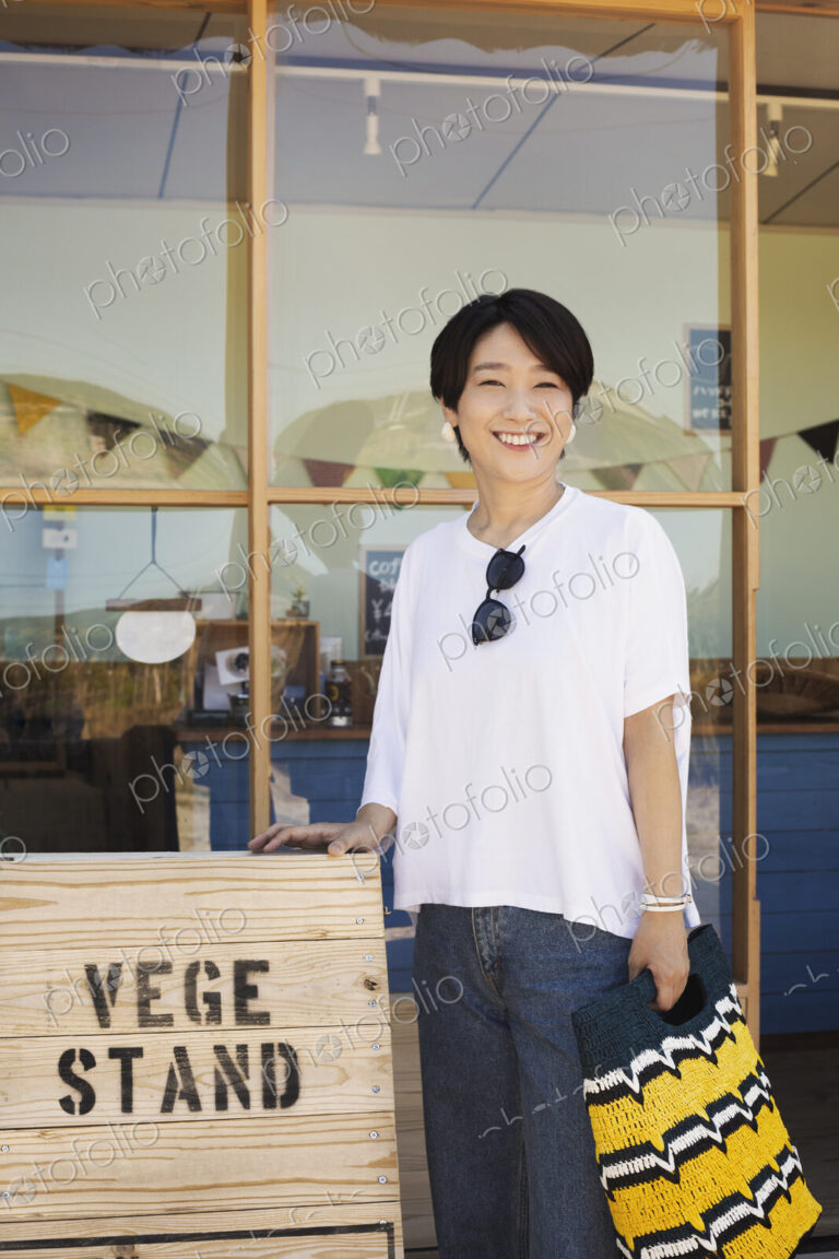 Japanese woman standing outside a farm shop, holding shopping bag, smiling at camera.
