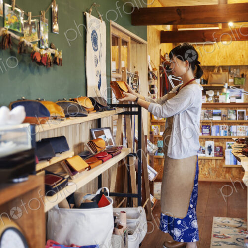 Japanese woman wearing apron standing in a leather shop, holding tan leather clutch bag. Japanese woman wearing apron standing in a leather shop, holding tan leather clutch bag.