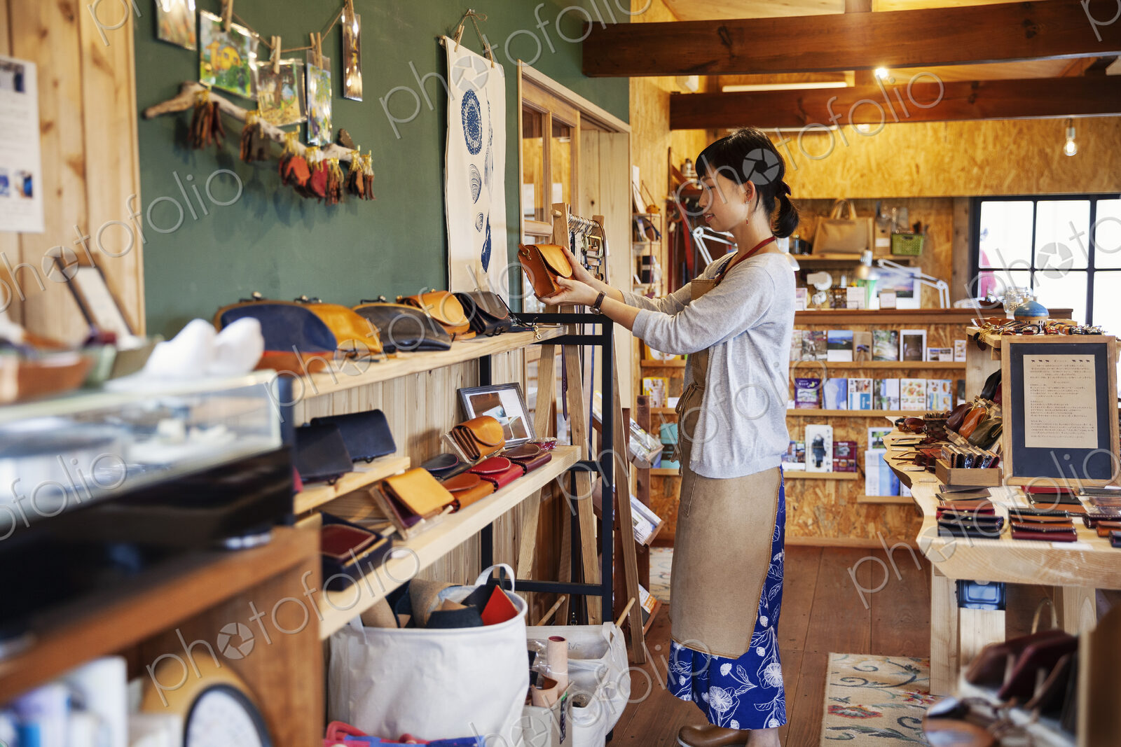 Japanese woman wearing apron standing in a leather shop, holding tan leather clutch bag.