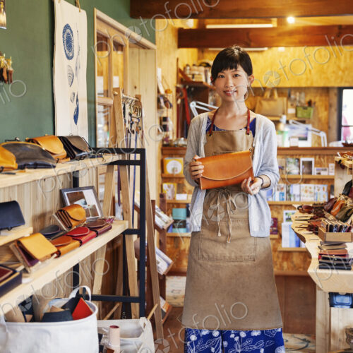 Japanese woman wearing apron standing in a leather shop, holding tan leather clutch… Japanese woman wearing apron standing in a leather shop, holding tan leather clutch…