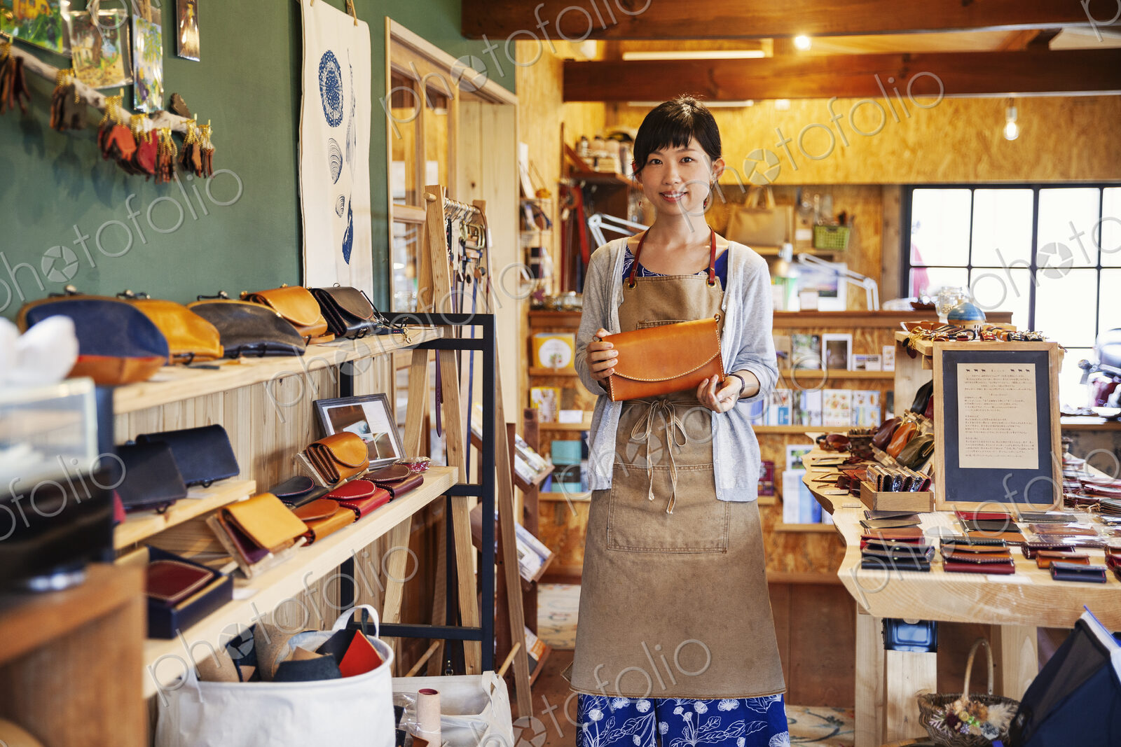 Japanese woman wearing apron standing in a leather shop, holding tan leather clutch…