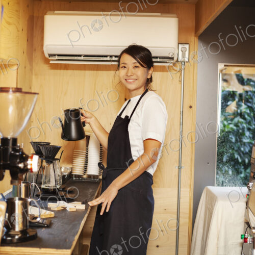 Japanese woman wearing apron standing in an Eco Cafe, preparing coffee, smiling at camera. Japanese woman wearing apron standing in an Eco Cafe, preparing coffee, smiling at camera.