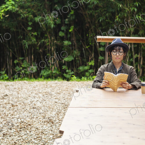 Japanese woman wearing glasses and hat sitting at a table outside Eco Cafe, reading book.