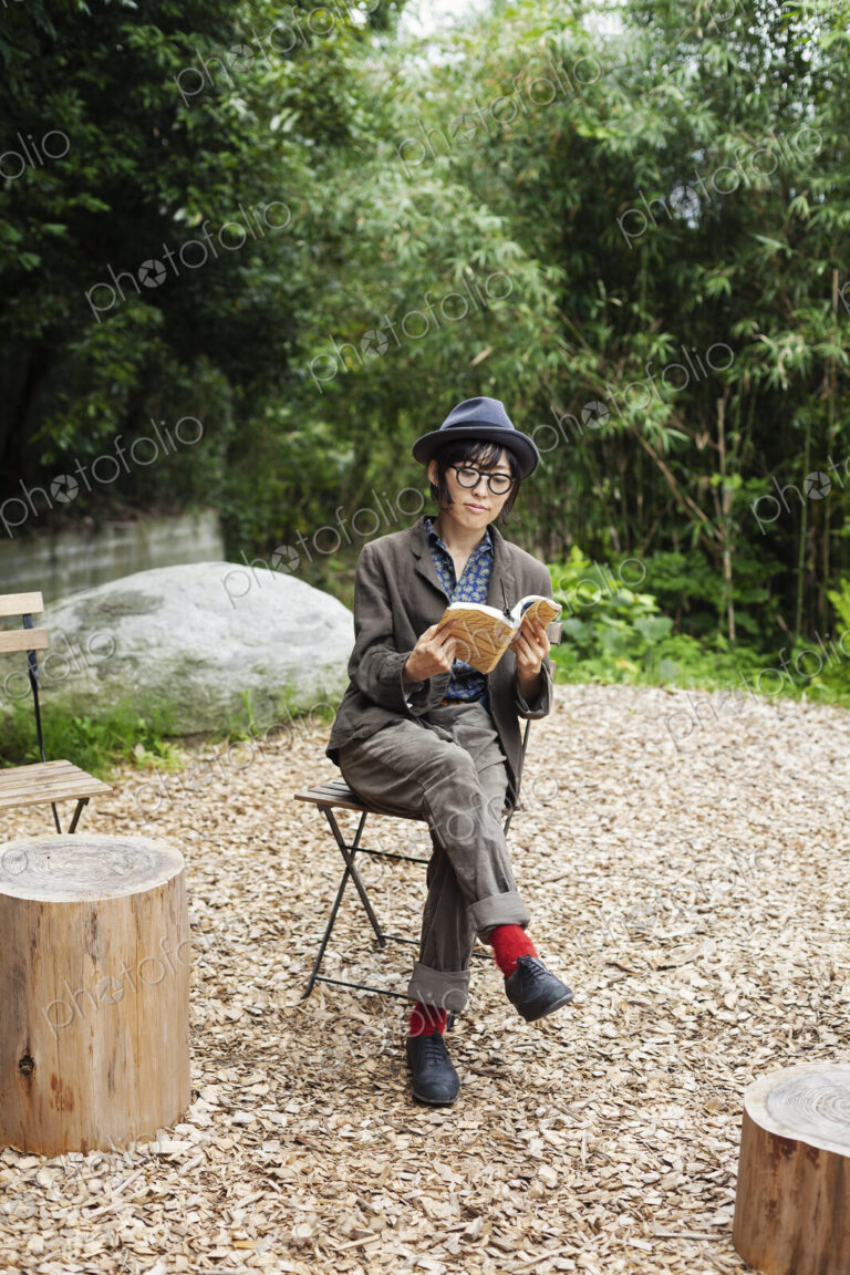 Japanese woman wearing glasses and hat sitting on chair outside Eco Cafe, reading book.