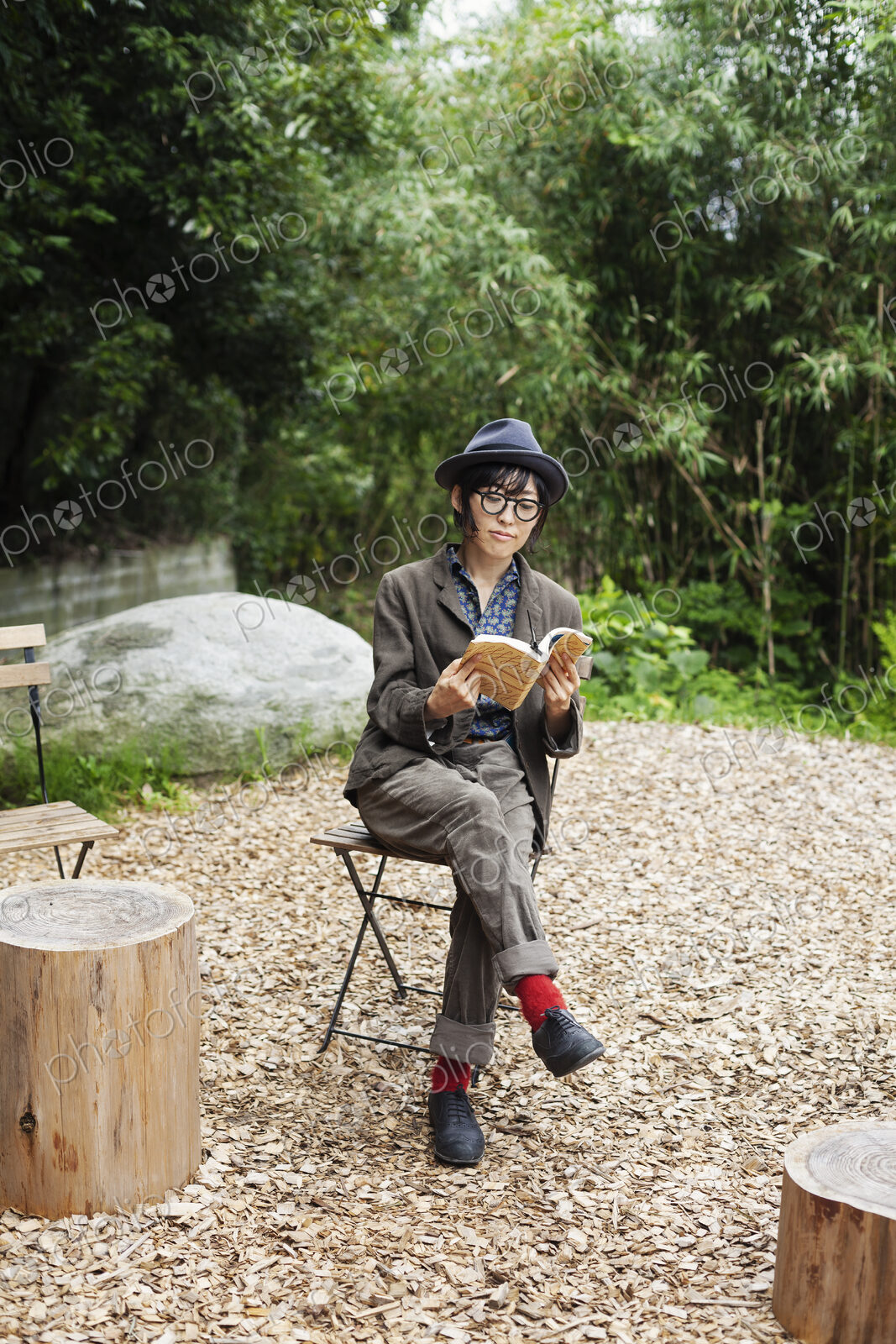 Japanese woman wearing glasses and hat sitting on chair outside Eco Cafe, reading book.