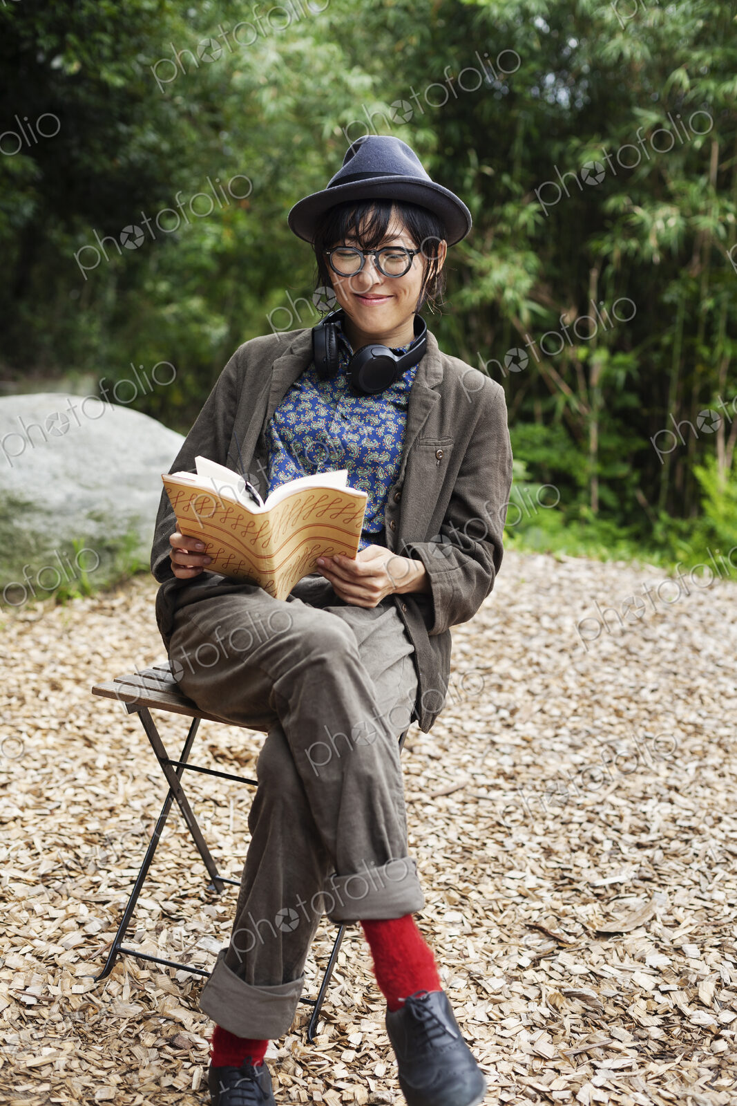 Japanese woman wearing glasses and hat sitting on chair outside Eco Cafe, reading book.