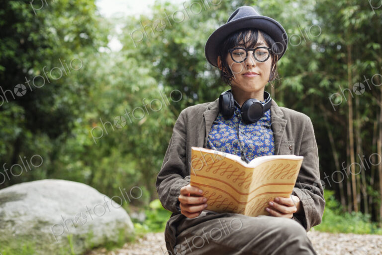 Japanese woman wearing glasses and hat sitting on chair outside Eco Cafe, reading book.