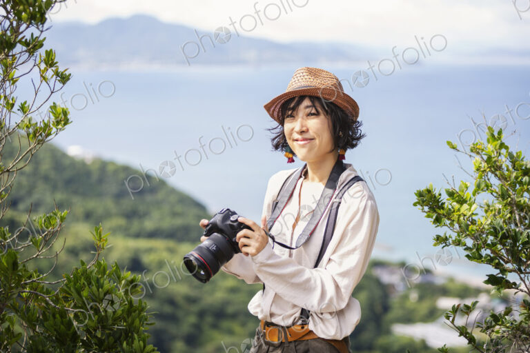 Japanese woman wearing hat and carrying backpack and camera standing on a cliff, ocean…