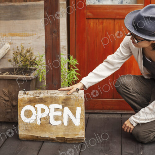 Japanese woman wearing hat and glasses kneeling in front of a leather shop, holding…