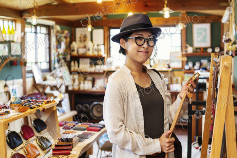 Japanese woman wearing hat and glasses standing in a leather shop, holding leather belt.