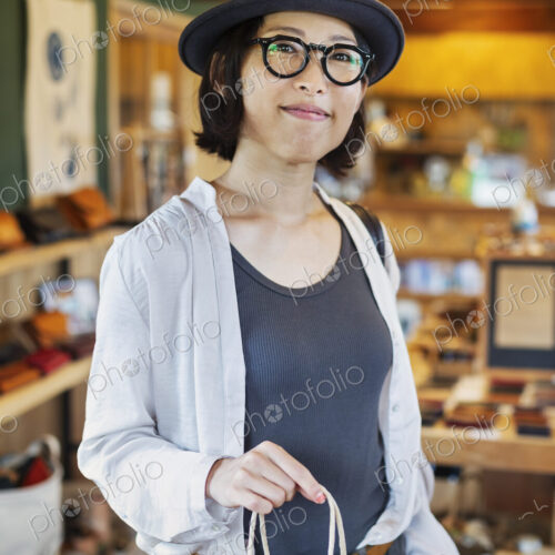 Japanese woman wearing hat and glasses standing in a leather shop, holding shopping… Japanese woman wearing hat and glasses standing in a leather shop, holding shopping…