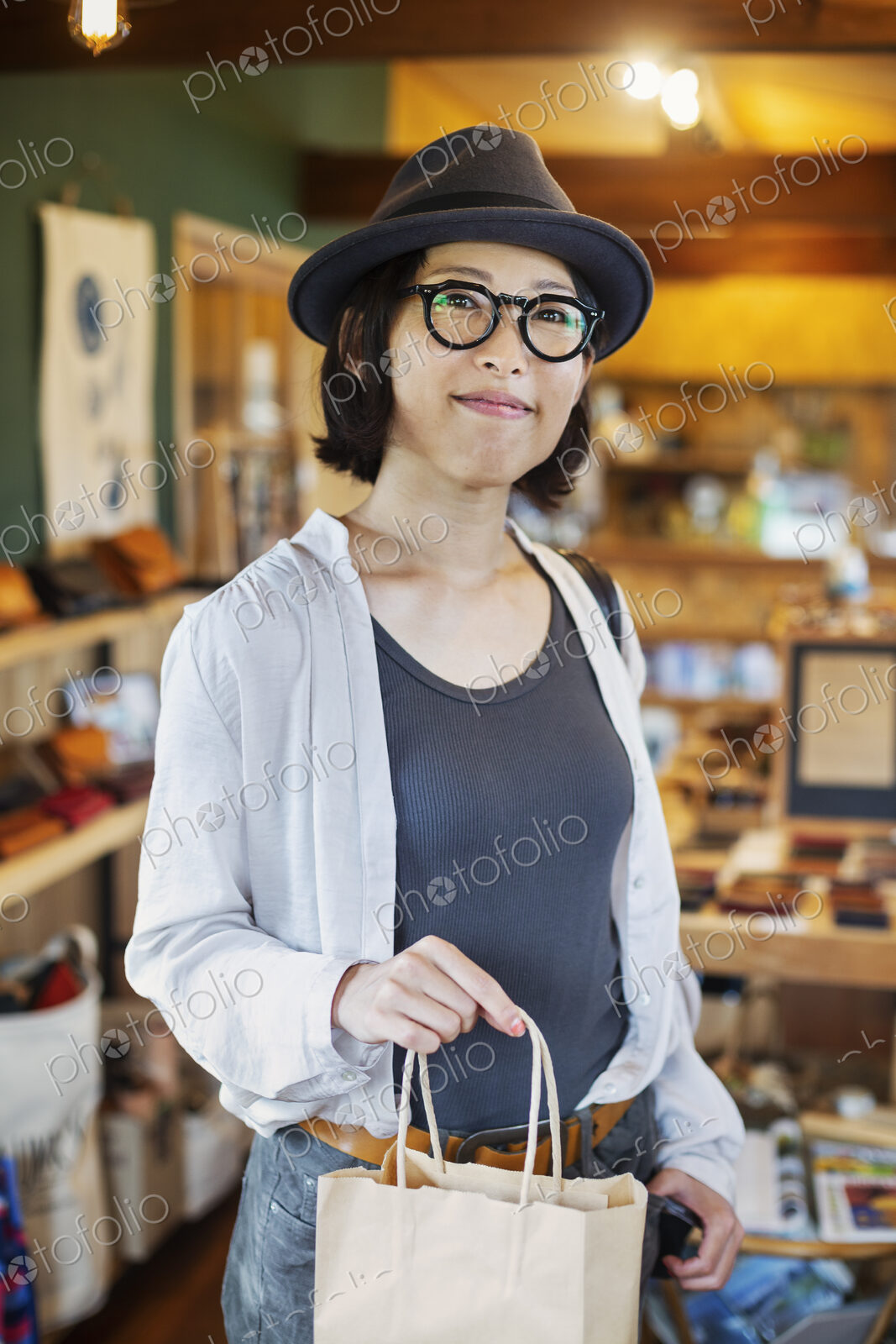 Japanese woman wearing hat and glasses standing in a leather shop, holding shopping…