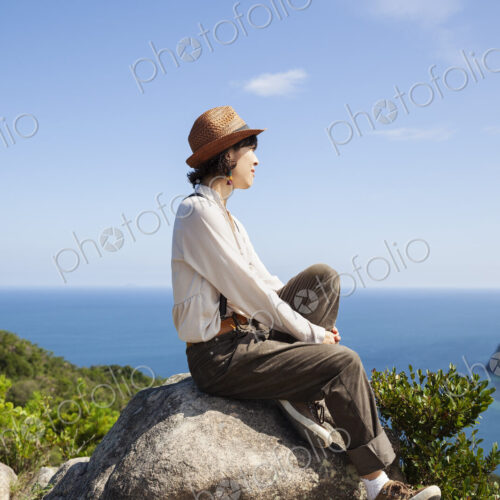 Japanese woman wearing hat sitting on a rock on cliff, ocean in the background. Japanese woman wearing hat sitting on a rock on cliff, ocean in the background.