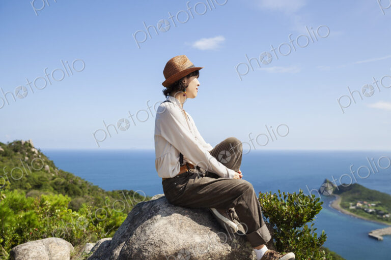 Japanese woman wearing hat sitting on a rock on cliff, ocean in the background.