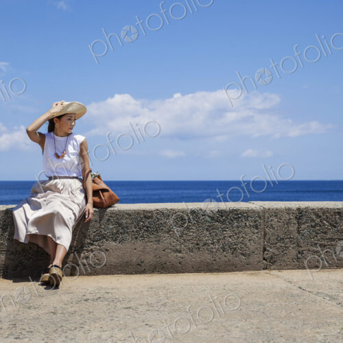 Japanese woman wearing hat sitting on a wall, ocean in the background. Japanese woman wearing hat sitting on a wall, ocean in the background.