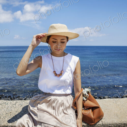 Japanese woman wearing hat sitting on a wall, ocean in the background.