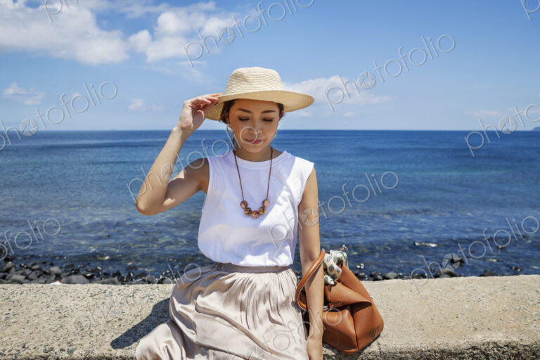 Japanese woman wearing hat sitting on a wall, ocean in the background.
