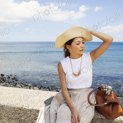 Japanese woman wearing hat sitting on a wall, ocean in the background.