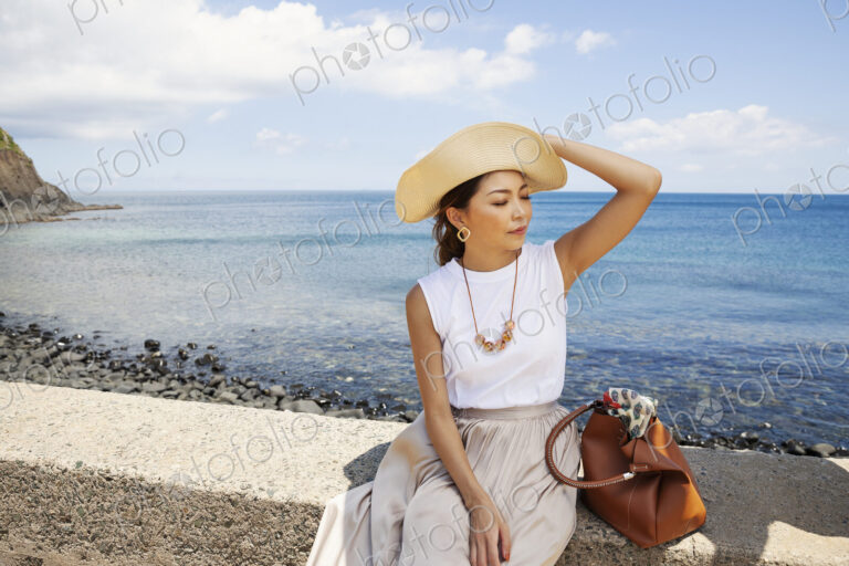 Japanese woman wearing hat sitting on a wall, ocean in the background.