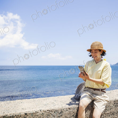 Japanese woman wearing hat sitting on a wall, ocean in the background.