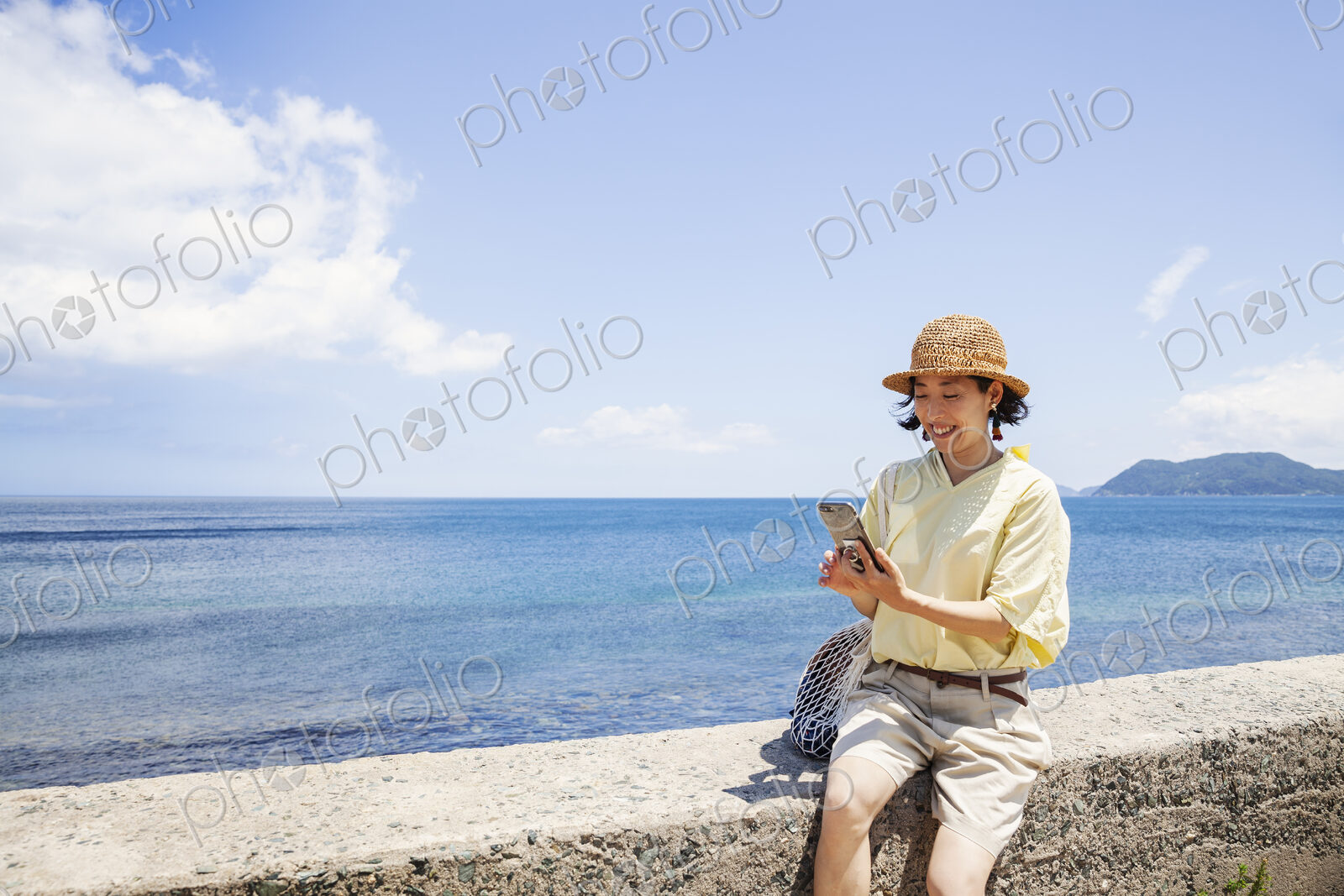 Japanese woman wearing hat sitting on a wall, ocean in the background.
