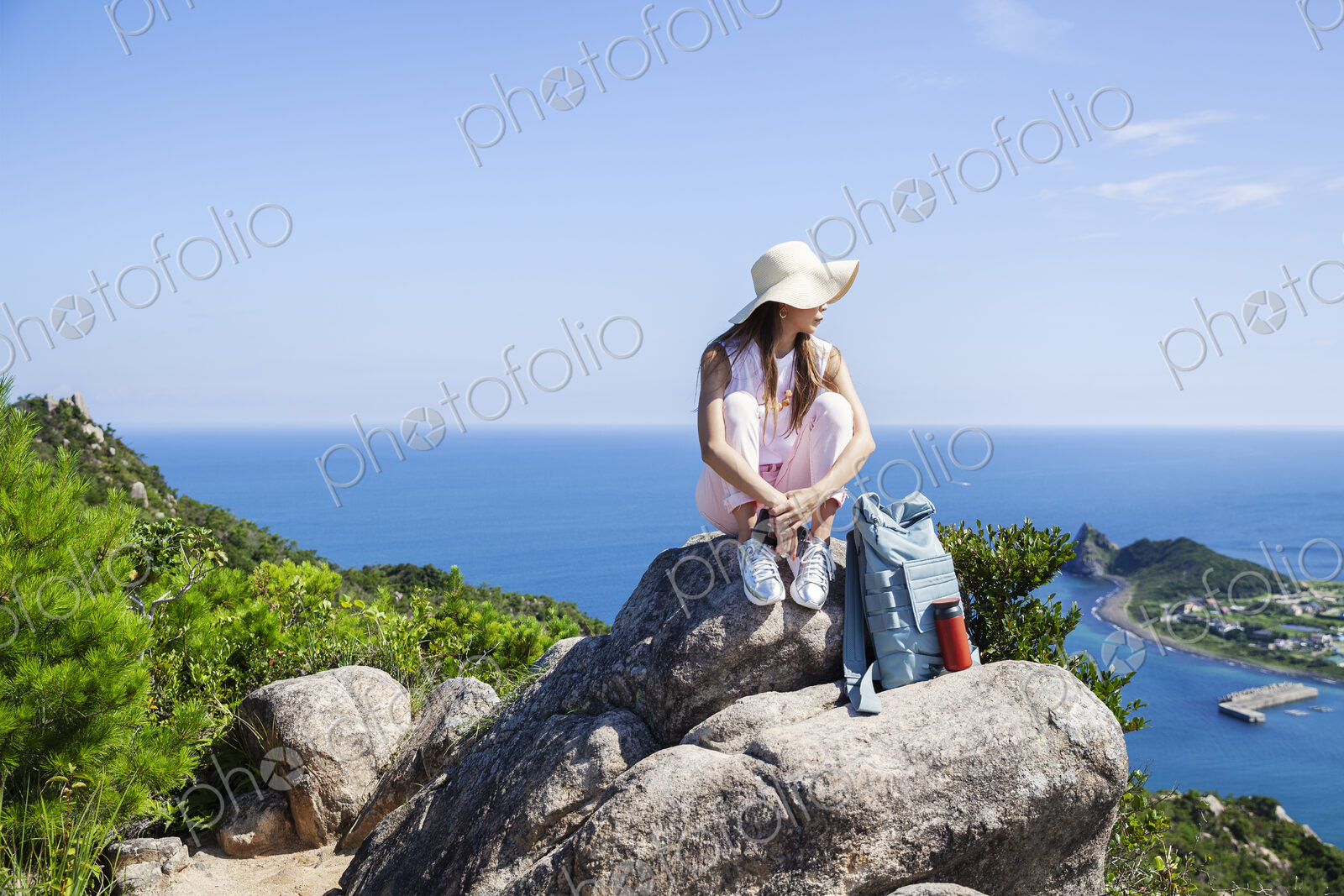 Japanese woman wearing hat sitting on rock on a cliff, ocean in the background.