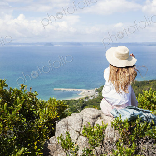 Japanese woman wearing hat sitting on rock on a cliff, ocean in the background. Japanese woman wearing hat sitting on rock on a cliff, ocean in the background.