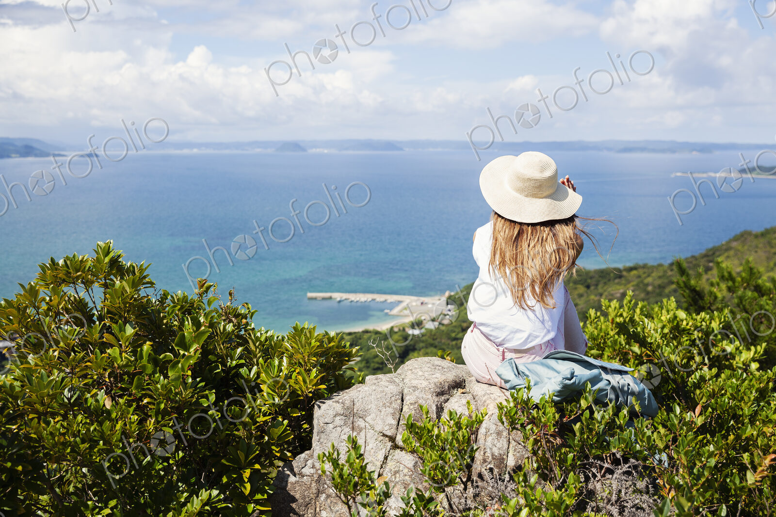Japanese woman wearing hat sitting on rock on a cliff, ocean in the background.