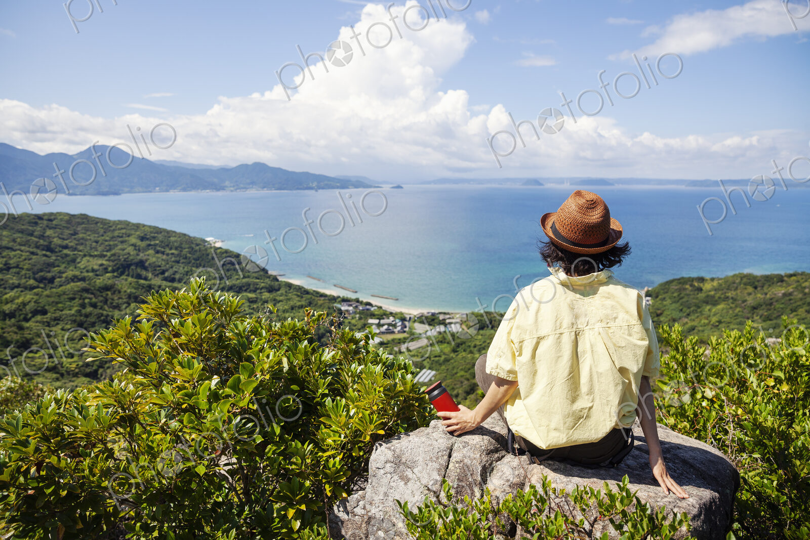 Japanese woman wearing hat sitting on rock on a cliff, ocean in the background.