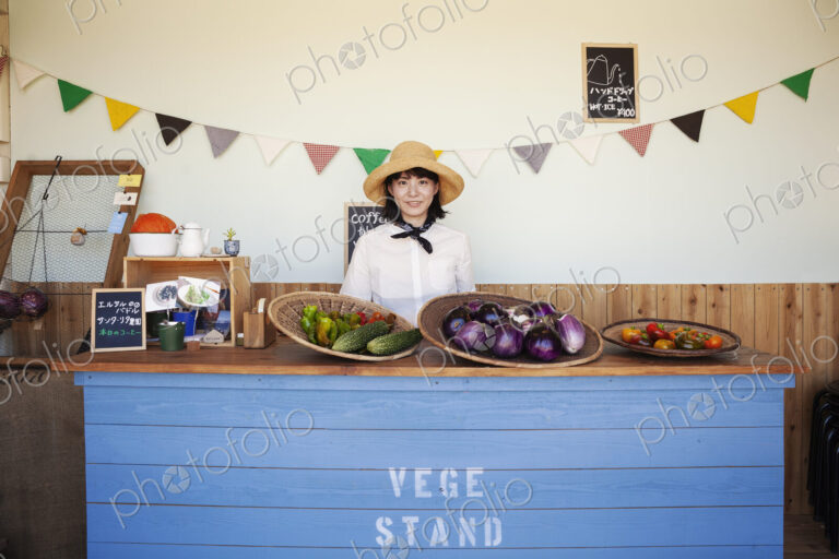 Japanese woman wearing hat standing behind counter in a farm shop, smiling at camera.