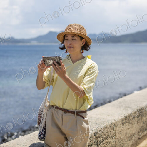 Japanese woman wearing hat standing by the ocean, taking picture with mobile phone. Japanese woman wearing hat standing by the ocean, taking picture with mobile phone.