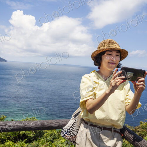 Japanese woman wearing hat standing on a cliff, taking selfie with mobile phone, ocean… Japanese woman wearing hat standing on a cliff, taking selfie with mobile phone, ocean…