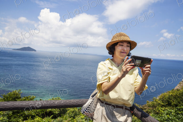 Japanese woman wearing hat standing on a cliff, taking selfie with mobile phone, ocean…