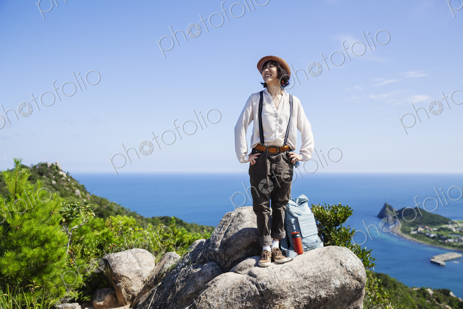 Japanese woman wearing hat standing on a rock on cliff, ocean in the background.