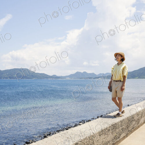 Japanese woman wearing hat standing on a wall, looking at ocean.