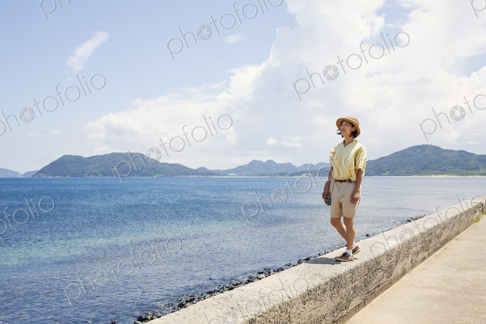 Japanese woman wearing hat standing on a wall, looking at ocean.