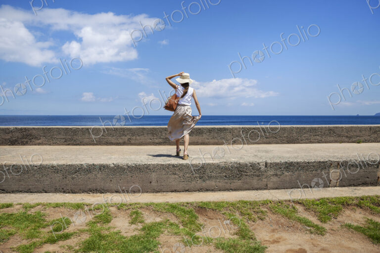 Japanese woman wearing hat standing on a wall, ocean in the background.