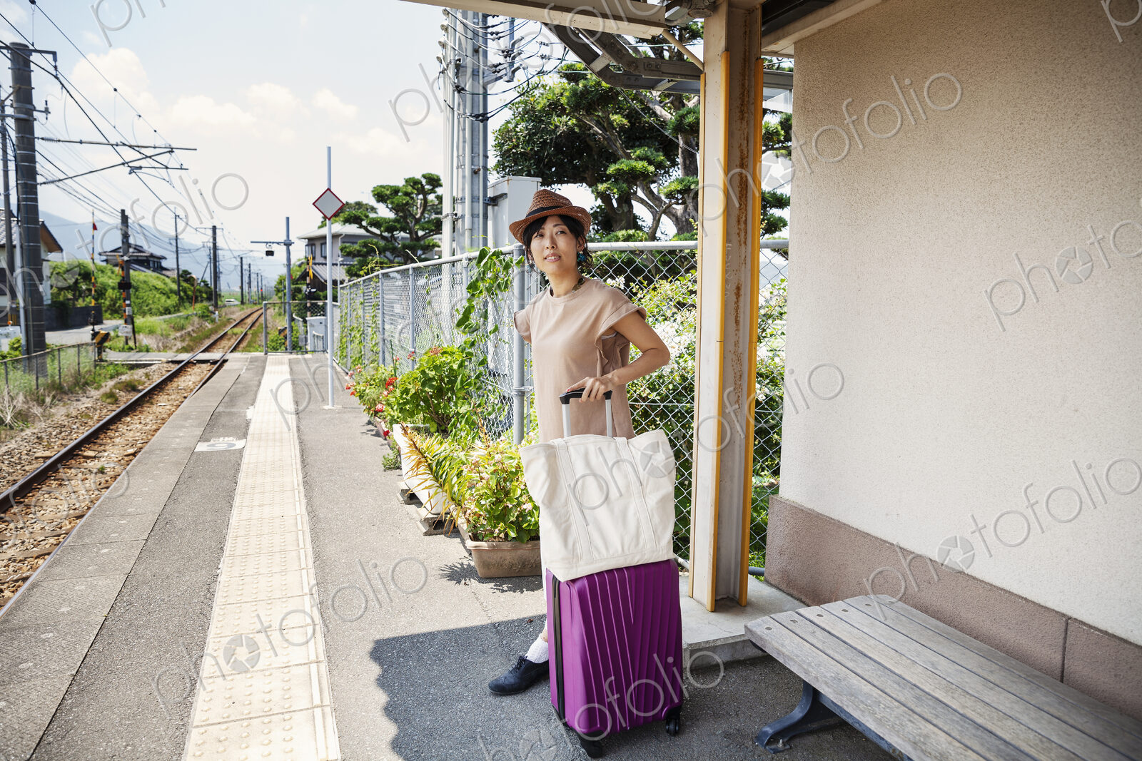 Japanese woman wearing hat standing on railway station platform with shopping bag and…