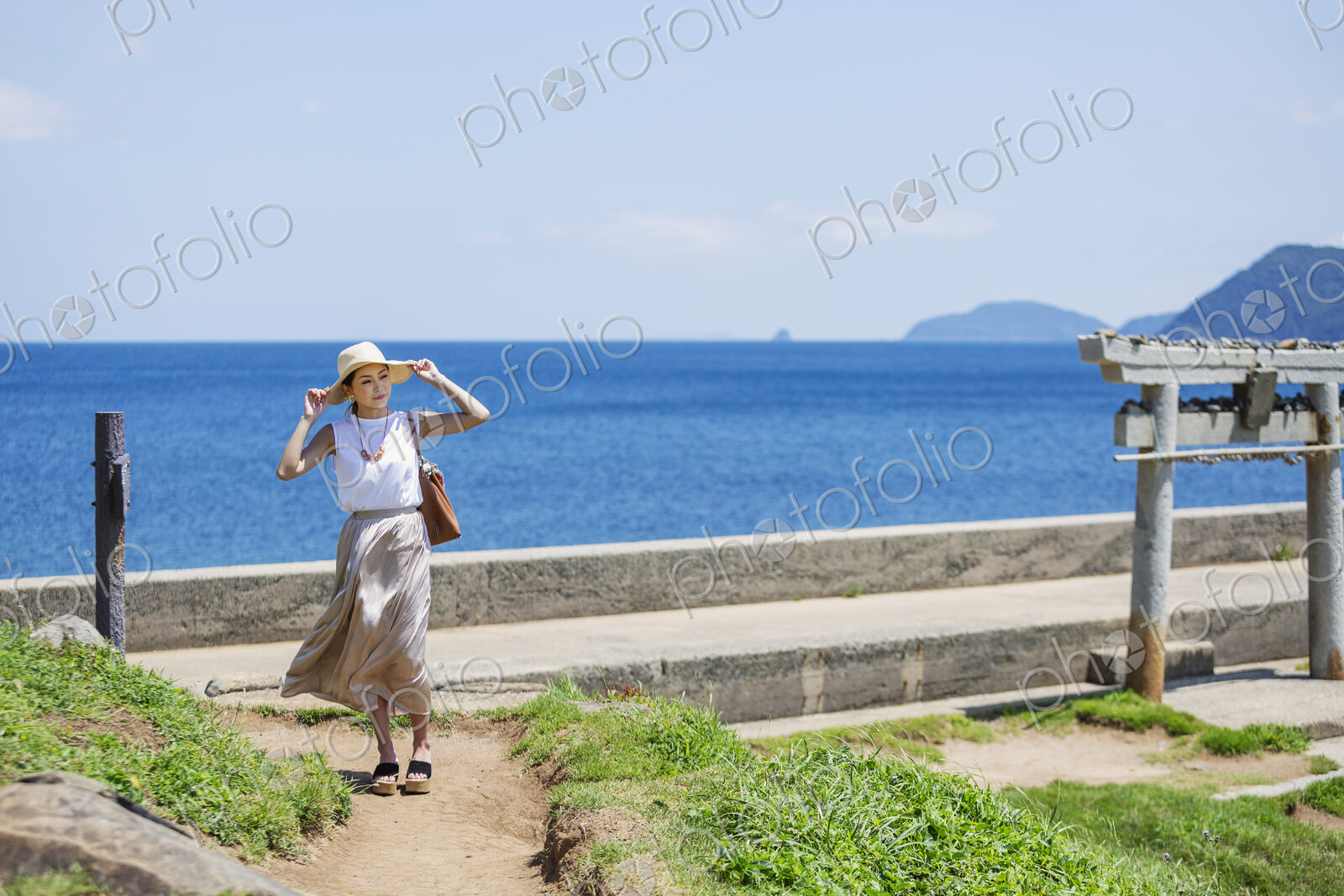 Japanese woman wearing hat walking along path by the ocean.