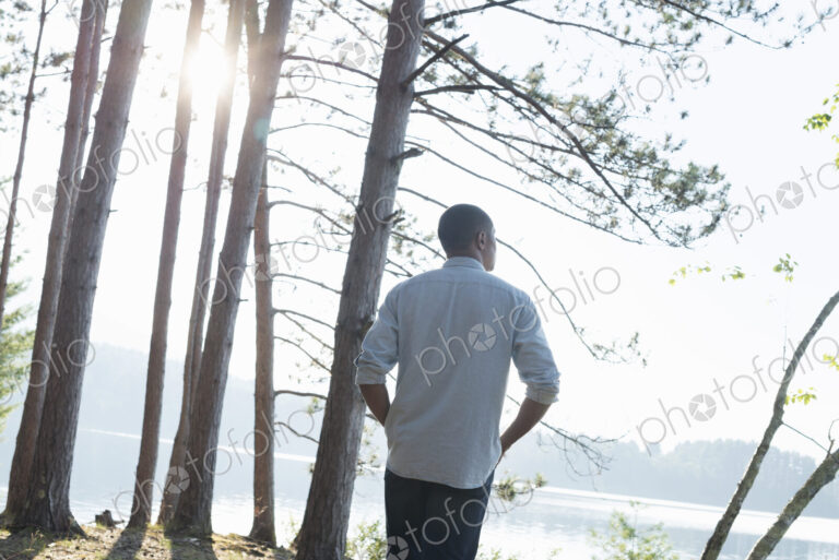 Lakeside. A man standing in the shade of pine trees in summer.