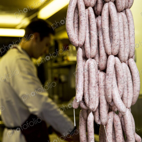 Large number of freshly made sausages hanging in a butcher shop, man wearing apron…