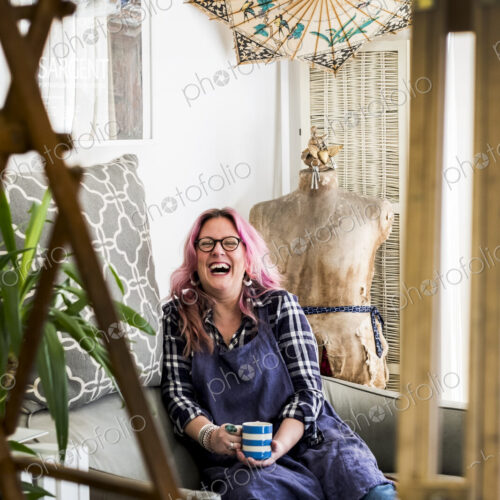 Laughing woman with long blond wavy hair with pink streaks sitting on a sofa, looking…