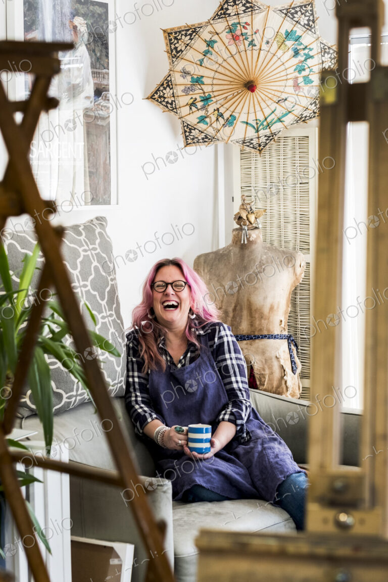 Laughing woman with long blond wavy hair with pink streaks sitting on a sofa, looking…
