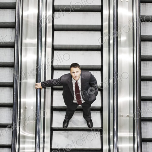 Looking down on a caucasian businessman looking up while riding an escalator in a… Looking down on a caucasian businessman looking up while riding an escalator in a…