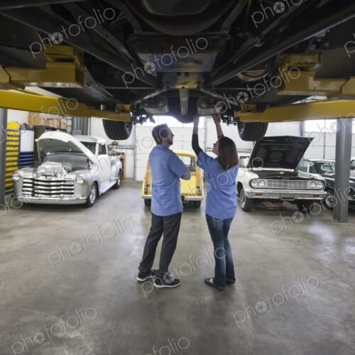 Male and female mechanics working on the underside of a car on a lift in a repair shop.