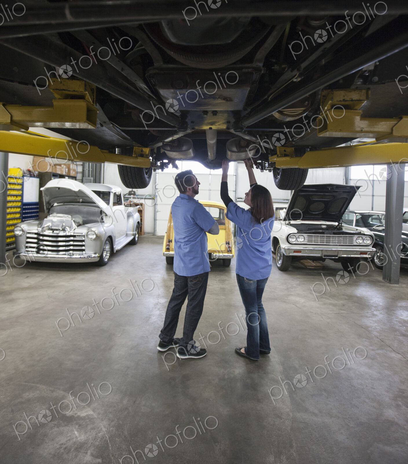 Male and female mechanics working on the underside of a car on a lift in a repair shop.