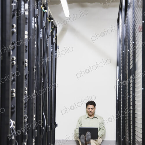 Male computer technician working on a lap top while sitting in an aisle of servers on… Male computer technician working on a lap top while sitting in an aisle of servers on…