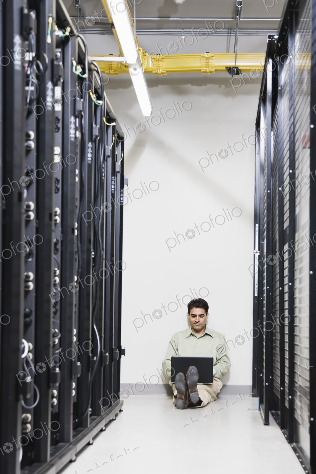 Male computer technician working on a lap top while sitting in an aisle of servers on…