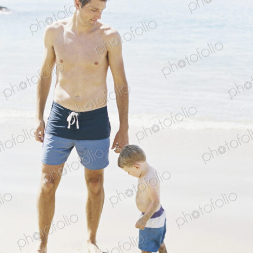 Man and his son wearing swimming trunks standing on a sandy beach by the ocean.